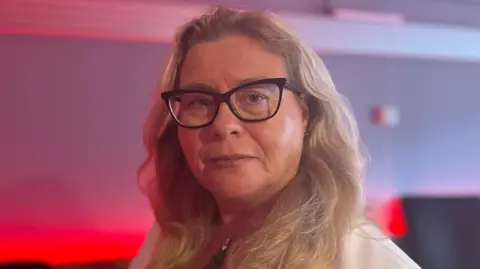 BBC A head and shoulders shot of Marcella Leonard. She has longer, blonde hair and is wearing black-framed cat eye glasses. She is wearing a white blazer and a black shirt. She is looking down the camera lens with a neutral expression on her face.