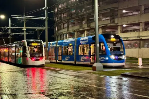 Graham Christie Two trams one white, the other blue, are parked in a street. The ground is wet and they are illuminated in the dark by street lights.