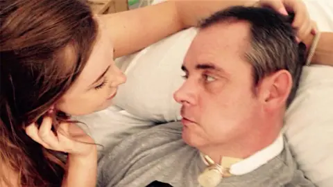 Simon Dobbin lying in a hospital bed, looking his daughter in the eye. She is holding his head and leaning towards him.