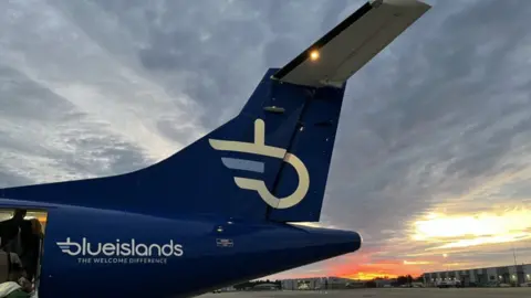 BBC The tail of a Blue Islands plane is visible on Jersey's airport runway. Passengers are boarding the plane and a sunset is seen in the background. 