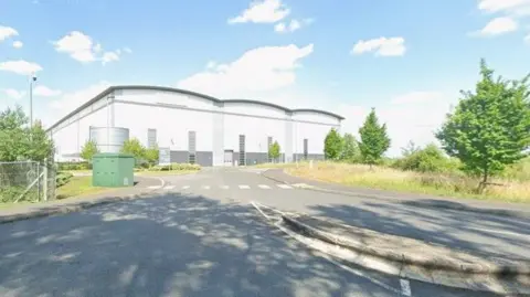 Google Large white and grey warehouse with road leading up to it and tree lined verges either side.