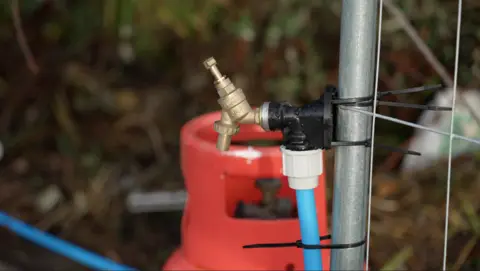 BBC A tap on the meanwhile site. It is attached to a grey metal fence post with black cable ties