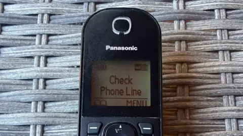Vikki Irwin/BBC The top of a landline telephone which displays the message "check phone line" on its small square screen. 
