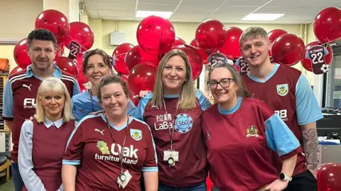 A mixed group of men and women wear a range of Burnley United Football Club's shirts - dark red body, light blue sleeves - or red and blue clothing. They stand in front of a group of red balloons, and bunting of small Burnley United football shirts. 