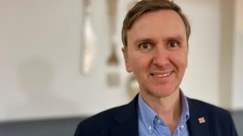 Andrew Pakes stares at the camera. He wears a navy jacket and a light blue shirt. 