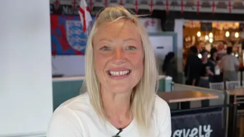 Helen Foster. A woman with long, blonde hair looking at the camera. She is wearing a white shirt and sitting in a pub that has England flags up.