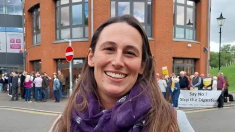 Nadia Lincoln/LDRS A head and shoulders image of Laura Weston. She is smiling into the camera and standing in front of a large group of what appears to be protesters positioned in front of a building. 