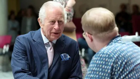 King Charles, wearing a pinstripe blue suit and pink tie, talks to a man (seen from behind) in a patterned blue shirt and glasses.