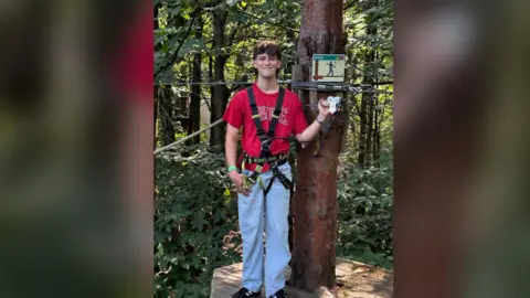 Family Handout Hamish Wilson - a man with a red top and blue jean on, underneath a harness, standing next to a tree