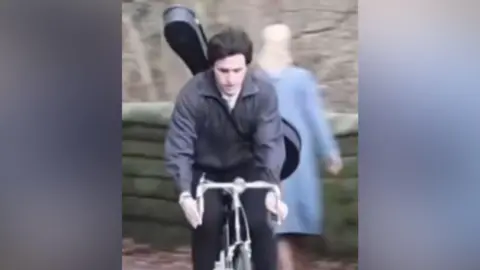 Actor Paul Mescal in character as Paul McCartney riding a bike with a guitar on his back past a brick wall
