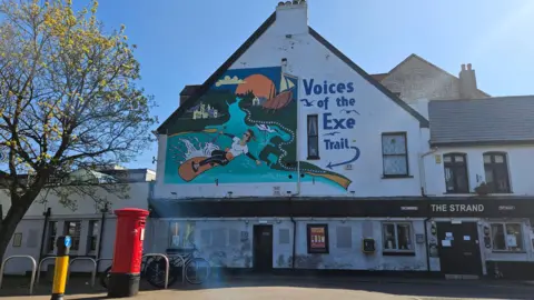 A general view of the Strand Inn pub. There is a red post box outside the pub. On the wall of the front of the pub there is a mural which says "Voices of the Exe Trail" with a blue arrow pointed at a painting of a woman surfing. There is a boat on the land in the painting and on the opposite side of the river a white church building. The pub says "The Strand" on it. 