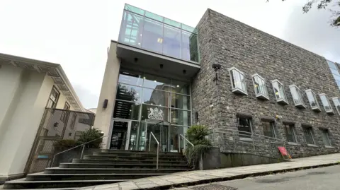 BBC Guernsey’s Royal Court building with modern glass balconies and traditional stone facade rises beside a sloping street, featuring steps, railings, tall windows, nearby trees, and bright daylight urban surroundings quiet.