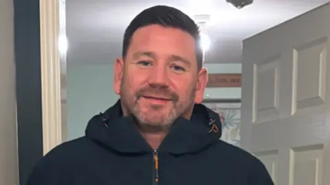 Russell Stamp. He has brown hair and stubble on his chin. He is wearing a hooded coat and is standing in a doorway.