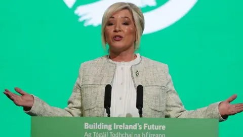 PA Media A woman with blonde hair, a grey jacket and white top stands behind a lectern with two black microphones. There is a green and white screen in the background.