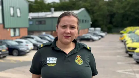 Welsh Ambulance Service Emily Hilton wearing her dark green paramedic uniform of polo shirt, trousers and belt with badge attached. She has long dark hair tied back in a pony tail and poses with her hands behind her back next to a row of parked ambulances on one side and cars on the other