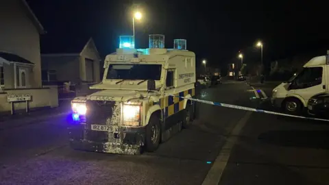 A police vehicle sits in the middle of a road. It is dark. The large vehicle is white. A police cordon tape is across its bonnet. A road sign reading Tullymore Road is to its left