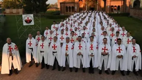 Brother John Michael A large group of men stand in formation wearing white robes with red crosses on their chests. They are in front of a historic brick church, with a lawn and a banner displaying a similar cross emblem.