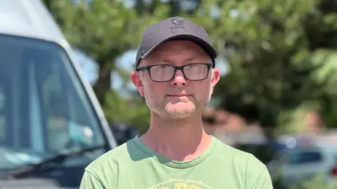 Man in his early 50s in a lime green t-shirt and a Liverpool FC baseball cap. He wears black rimmed glasses.