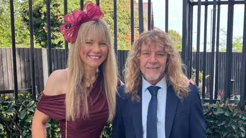 Supplied A woman and a man dressed smartly standing in front of a black fence. The woman on the left has a maroon dress and has long blonde hair with a maroon bow pinned to her head. The man has long curly blonde hair, a dark blue suit and tie and a light blue shirt. Behind them is another wooden fence further in the background, a house and many green trees.