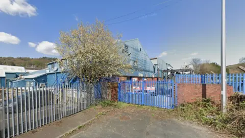 Google An industrial area. From a public footpath, you can see a warehouse building which houses the ironworks factory. The metal gates onto the ground are painted blue.