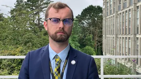 A person wearing glasses, a suit and a patterned tie stands in front of a modern concrete building with rows of windows, with trees and greenery behind.