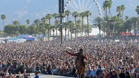Getty Images Singer and rapper T-Pain performing at the Coachella Stage with crowds waving their hands in the air and palm trees visible in the background as well as a large ferris wheel and marquees and mountains behind