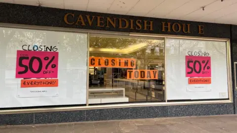 The shop window of Cavendish House saying 'closing today'. Much of the window is covered by large pieces of white paper, and there are posters saying "50% off" in black text on a pink background