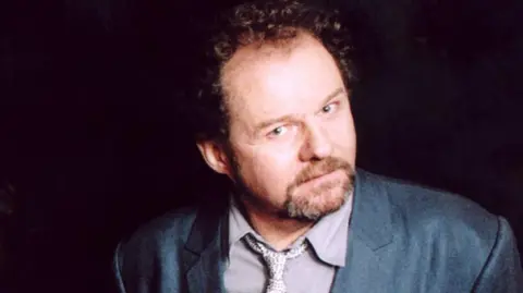 A head-and-shoulders portrait of Mike Figgis. He is looking up to the camera. He has curly dark hair and a beard. He wears a dark grey suit with a pale lilac shirt and tie.