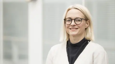A blonde woman with glasses looks at the camera and smiles. She is wearing a black top and a white cardigan, and is standing in front of a grey background.