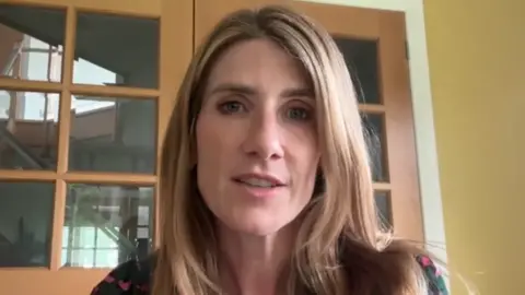 A woman, Kirsty Watson, with long hair, in a room in a house looking at the camera.
