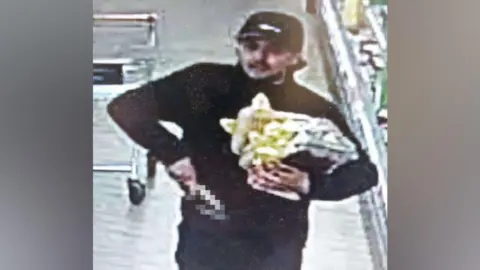 Gloucestershire Constabulary Grainy CCTV still of a man wearing a black cap and black clothes walking through a supermarket. He has a knife in his hand and he is carrying several packets of steak.