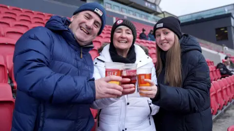 PA Media Three fans smile at the camera as they touch their pint glasses together in the stands at Ashton Gate before a game involving Bristol City Women's FC. Two of the group are women and one is a man and they are all dressed in winter clothing