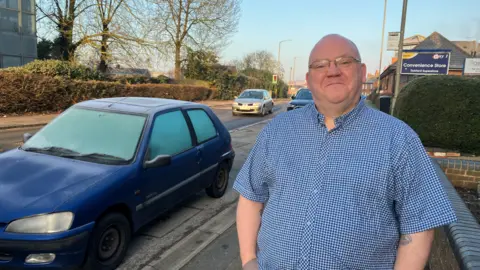 Amy Holmes/BBC A picture of a man, standing on a pavement next to a road and a blue car. He has his hands in his pocket and is wearing a blue short sleeved shirt. He has glasses on.