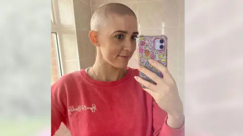 Handout Vicky Green smiles as she takes a selfie. She has lost her hair and is wearing a pink top and is holding a phone with a decorative phone case featuring images such as smiley faces, hearts and flowers.
