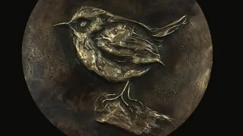 ALICE DUDLEY A round piece of gold-coloured metal with a bird engraved in it. The backgrounder is much darker.