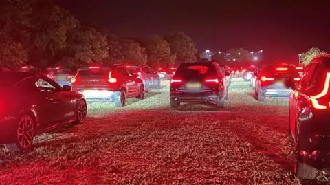 A long line of cars queueing up on a field. It is dark. The cars' tail-lights are shining red.