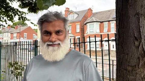 Nadim is stood behind a fence and next to a tree. He is wearing a grey t-shirt. 
