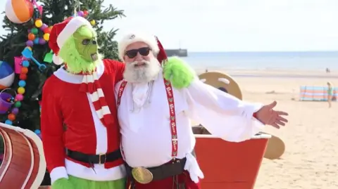 A man dressed as Santa, with a red and white Christmas hat, sunglasses, a white shirt, red braces and a big belt with a gold round buckle, puts his arm round a man dressed as the Grinch, with green fur and a red hat, jumper and scarf. They are standing on a golden beach on a bright summer's day with the blue sea in the background. A Christmas tree and a red sleigh are behind them.