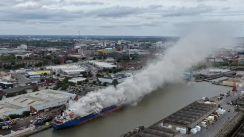 Dale Baxter/BBC A blue and red ship in a dock which has lots of grey smoke billowing out from a fire. The Hull skyline is visible in the distance.
