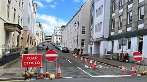 Road closed and no entry signs at mouth of a street featuring office buildings with parked cars and pedestrians at the top end