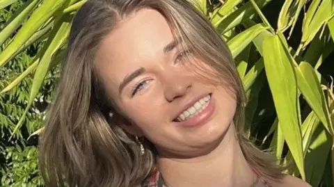 Holly Bowes smiles for the camera outside. There are many green plants behind her.