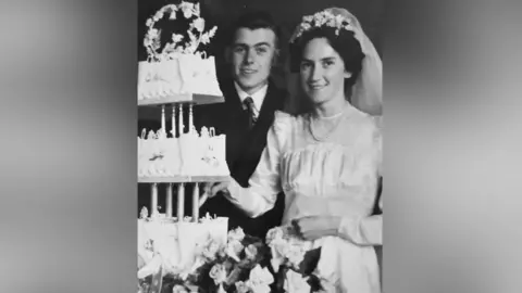 Family photo Black and white photograph, dating from 1950, of a couple on their wedding day. She is wearing a high necked white dress with long sleeves, and a veil pulled back over long dark hair. They are standing behind a large stand of roses and a two-tiered wedding cake, which she is about to cut.
