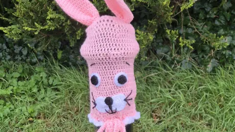 A crocheted pink bollard cover in the shape of a bunny face. It has two eyes, a nose and whiskers. Pink crocheted ears are on top of the bollard. Bushes are behind the bollard.