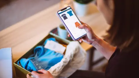 Getty Images Woman looks at smartphone showing clothing with a package on the table showing a blue top