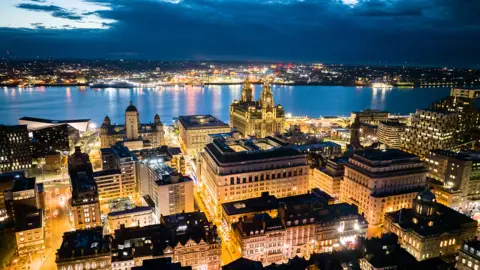 Liverpool City Council handout Aerial view of Liverpool city centre showing the three graces and the River Mersey with Wirral in the background. It is taken in the evening with all the building lit up.