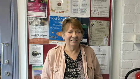 Ann Robb, who runs a line dancing class at Longtown Community Centre. She is standing in front of a notice board, wearing a pink jacket and black floral shirt.