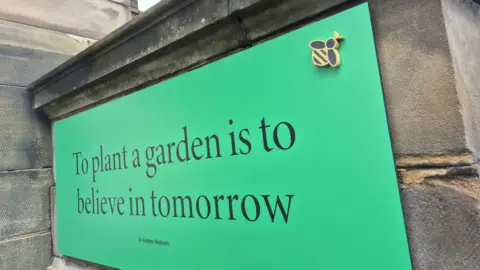 A large sign reading to plant a garden is to believe in tomorrow, with black lettering on a green background. A small model of a bee has been attached to the top right hand corner.