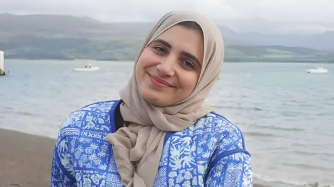 Jacob Lloyd/MFL Mentoring Head and torso shot of Firial Benamer, who is a woman of Libyan heritage in her early twenties. She is wearing a light tan headscarf tied in front of her neck and a light blue jacket with a pattern of different white leaves in a loose arrangement of squares across it. She is wearing light pink lipstick and has her head tilted to the side and is smiling directly at the camera. She is standing on a beach of either the sea or a lake, with hills in the distance and three small boats visible but slightly out of focus behind her.