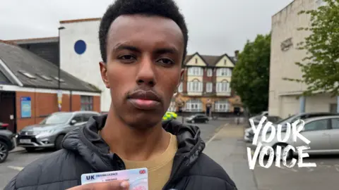 Robert is facing the camera and holding his drivers licence in his hand which he recently passed. He is standing on a residential street in Croydon with parked cars behind him.