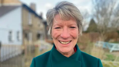 Pippa Heylings, the Liberal Democrat MP for South Cambridgeshire, is smiling at the camera. She has short, fair hair and is wearing a blue coat